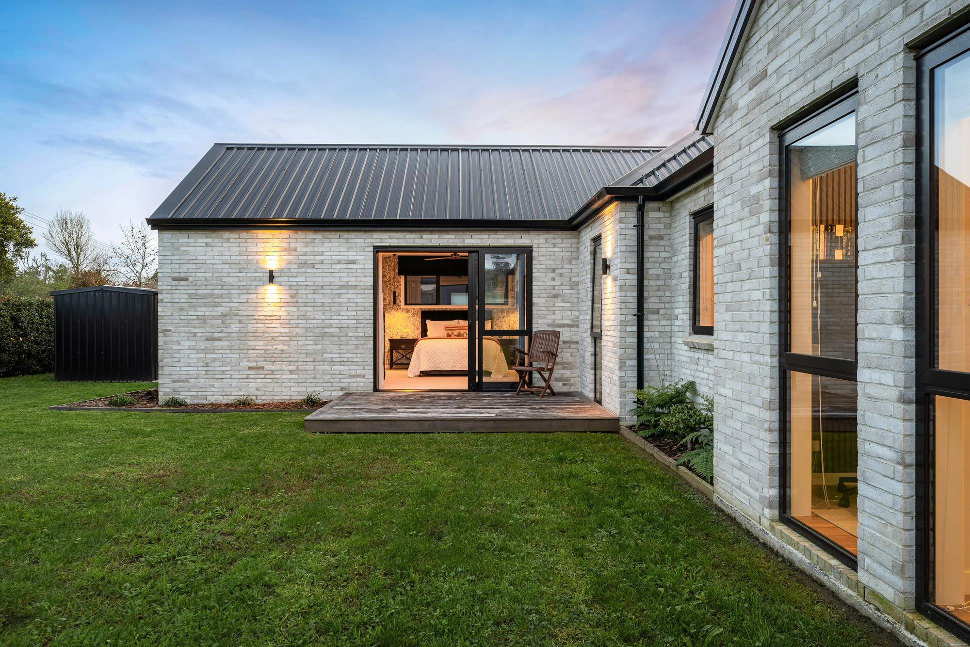 The Matakana home at dusk, white brick and a dark gable roof, a lit bedroom opening onto a timber deck and lawn