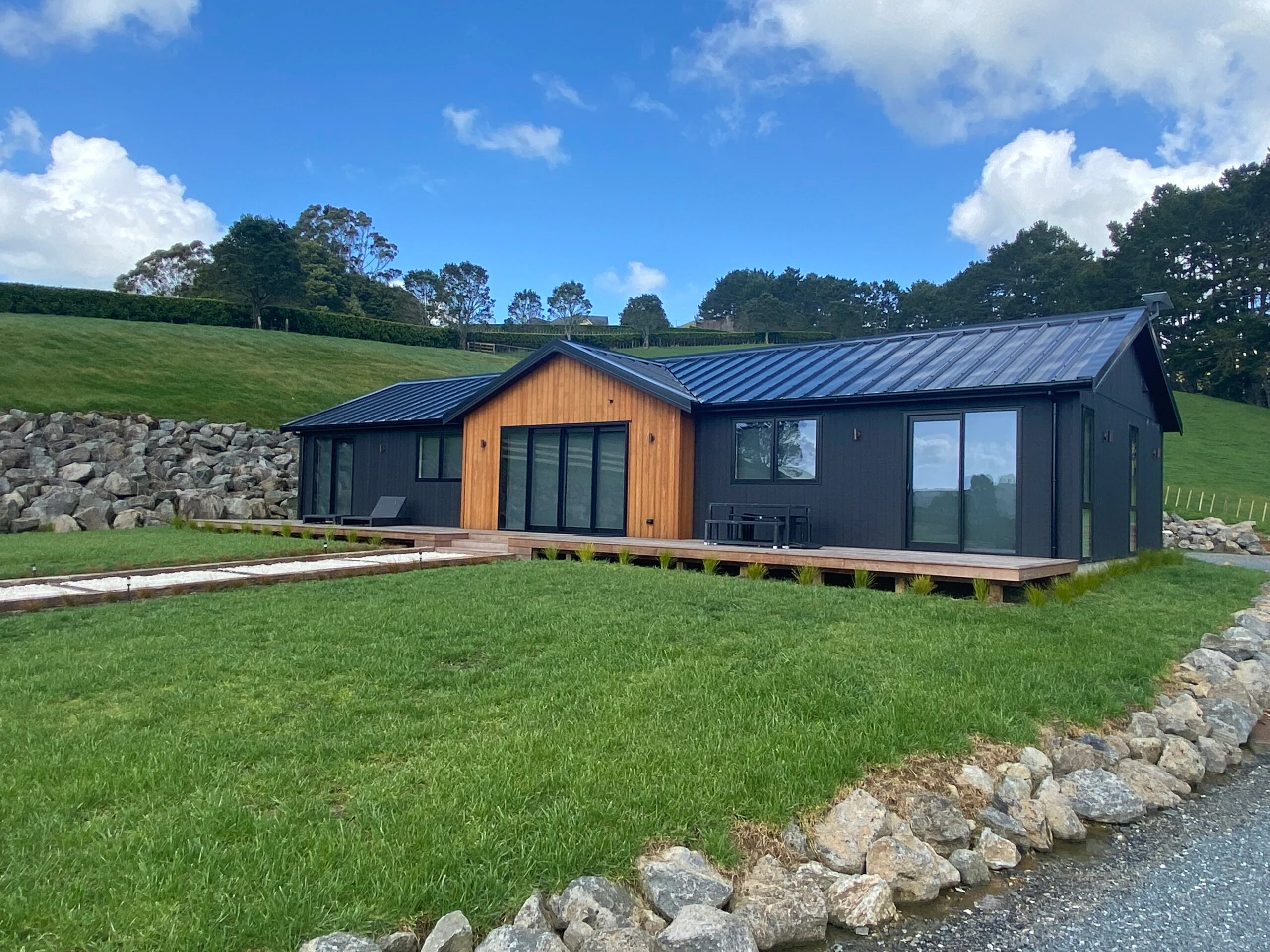 A dark-clad transportable home with a warm cedar gable entrance, set against green rural hills