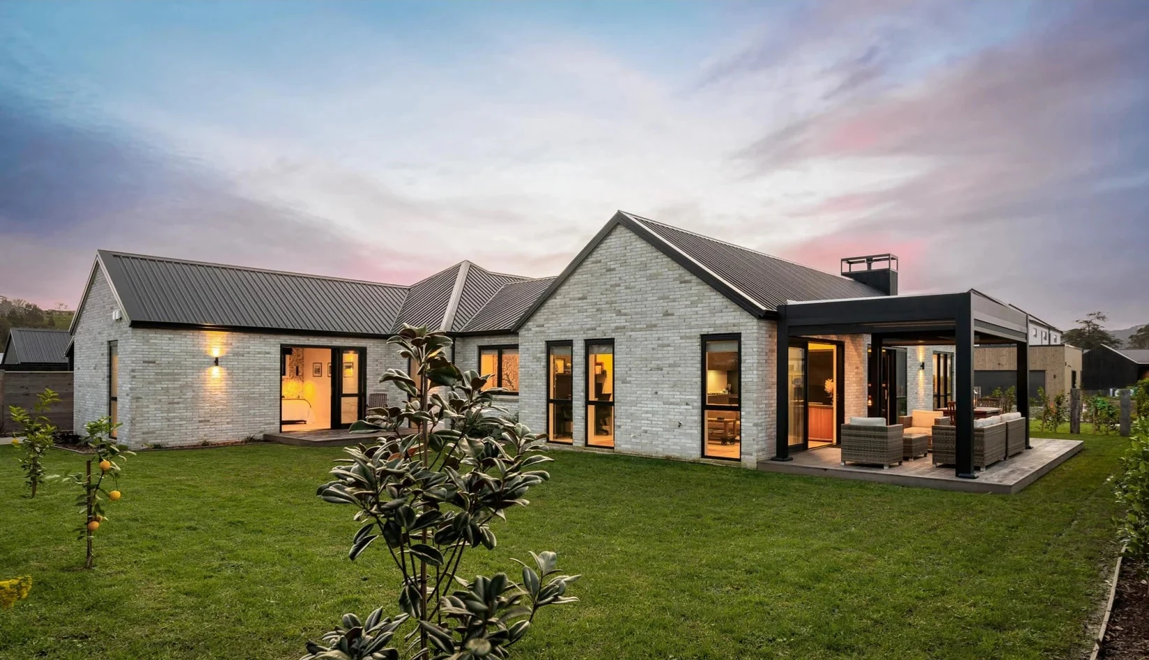 A pale brick gable home at dusk, warm light from inside reaching a lawn and a covered outdoor room